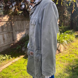 K&ouml;nnte beinhalten: Eine olivgr&uuml;ne Jacke mit mehreren Taschen und einer Knopfleiste. Die Jacke hat einen Kordelzug in der Taille und einen Kragen. Ein kleiner brauner Aufn&auml;her ist auf einer der Taschen sichtbar. Die Jacke wird auf einer Schaufensterpuppe pr&auml;sentiert.