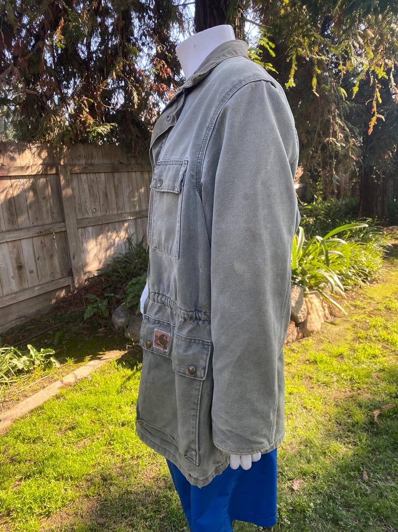 K&ouml;nnte beinhalten: Eine olivgr&uuml;ne Jacke mit mehreren Taschen und einer Knopfleiste. Die Jacke hat einen Kordelzug in der Taille und einen Kragen. Ein kleiner brauner Aufn&auml;her ist auf einer der Taschen sichtbar. Die Jacke wird auf einer Schaufensterpuppe pr&auml;sentiert.