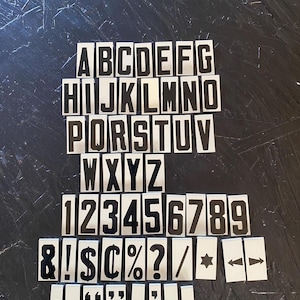 May include: A collection of black and white alphabet letters, numbers, and symbols. The letters A through Z are present, along with numbers 1 through 9, and various punctuation marks and symbols. The items are arranged on a dark, textured surface.