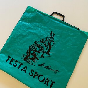 May include: A teal green plastic bag with a black handle and a black graphic of people playing sports. The bag has the words "TESTA SPORT" and "St. Moritz" printed in black.