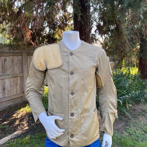 May include: Tan shooting jacket with padded shoulder and elbow patches. The jacket has a button-up front, two front pockets, and a tailored fit. The mannequin is wearing blue shorts. The jacket is displayed outdoors with a natural background.