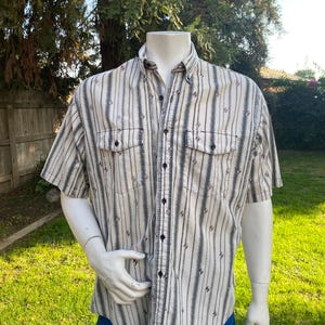 May include: Short-sleeved button-down shirt with vertical stripes in beige, gray, and black. The shirt has two chest pockets and a classic collar. The mannequin is wearing blue shorts, and the background is a grassy lawn.