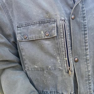 K&ouml;nnte beinhalten: Nahaufnahme einer abgenutzten, olivgr&uuml;nen Arbeitsjacke. Zu den Merkmalen geh&ouml;ren eine Brusttasche mit Rei&szlig;verschluss, zwei aufgesetzte Taschen mit Kn&ouml;pfen und ein durchgehender Rei&szlig;verschluss. Die Jacke weist Gebrauchsspuren mit Farbspritzern und messingfarbenen Kn&ouml;pfen auf.