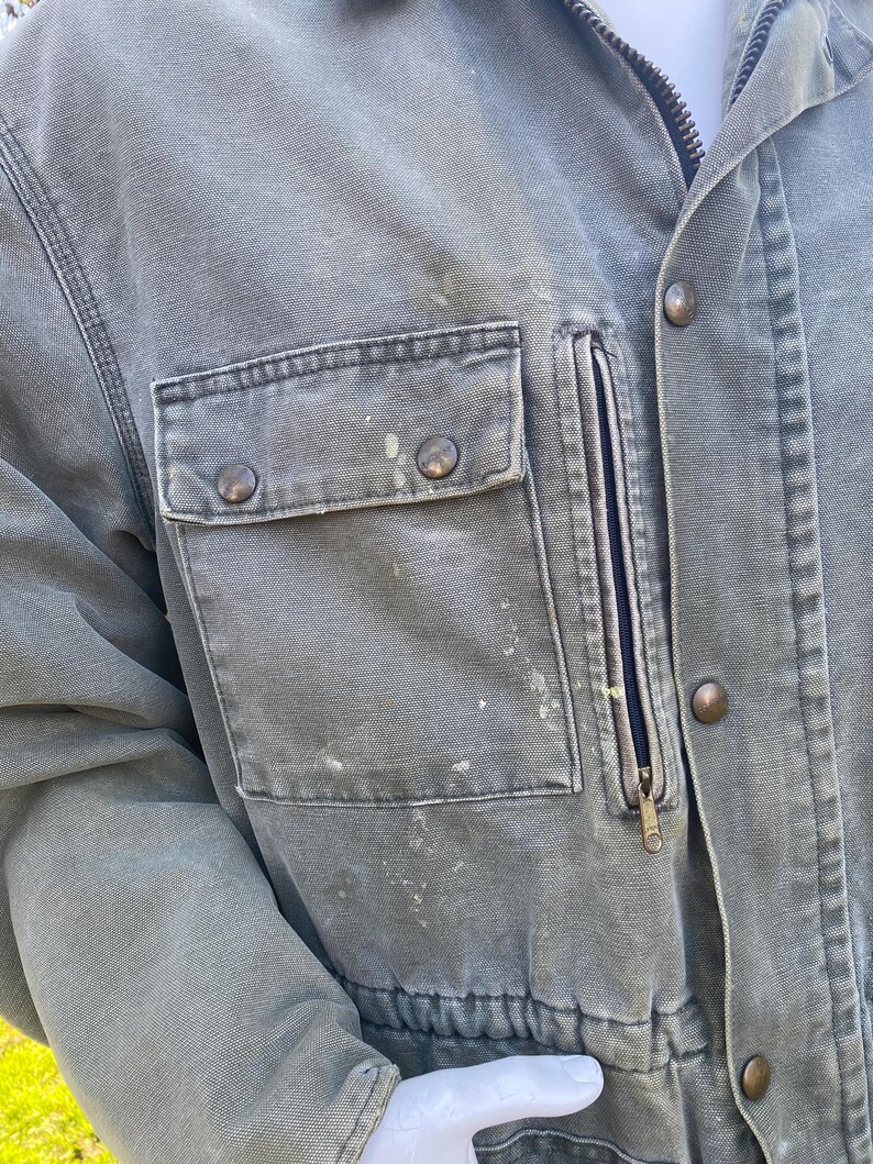 K&ouml;nnte beinhalten: Nahaufnahme einer abgenutzten, olivgr&uuml;nen Arbeitsjacke. Zu den Merkmalen geh&ouml;ren eine Brusttasche mit Rei&szlig;verschluss, zwei aufgesetzte Taschen mit Kn&ouml;pfen und ein durchgehender Rei&szlig;verschluss. Die Jacke weist Gebrauchsspuren mit Farbspritzern und messingfarbenen Kn&ouml;pfen auf.