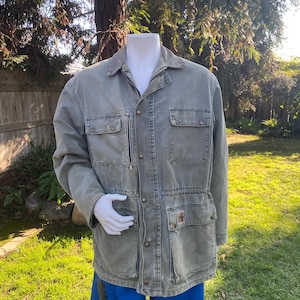 K&ouml;nnte beinhalten: Verblasste gr&uuml;ne Arbeitsjacke mit mehreren Taschen, Knopfleiste und Rei&szlig;verschlusstasche auf der Brust. Die Jacke hat einen Kragen und wird &uuml;ber blauen Shorts getragen. Das Carhartt-Logo ist auf einer der Taschen sichtbar.