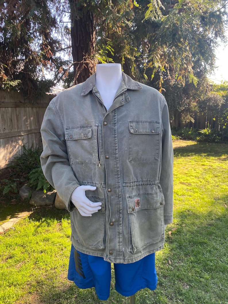 K&ouml;nnte beinhalten: Verblasste gr&uuml;ne Arbeitsjacke mit mehreren Taschen, Knopfleiste und Rei&szlig;verschlusstasche auf der Brust. Die Jacke hat einen Kragen und wird &uuml;ber blauen Shorts getragen. Das Carhartt-Logo ist auf einer der Taschen sichtbar.