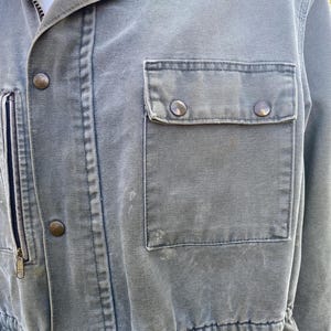 K&ouml;nnte beinhalten: Nahaufnahme einer gr&uuml;nen Canvas-Jacke mit Rei&szlig;verschluss und Druckkn&ouml;pfen. Die Jacke hat eine Brusttasche mit Druckkn&ouml;pfen und eine Taille mit Kordelzug. Der Stoff hat ein leicht abgenutztes Aussehen.