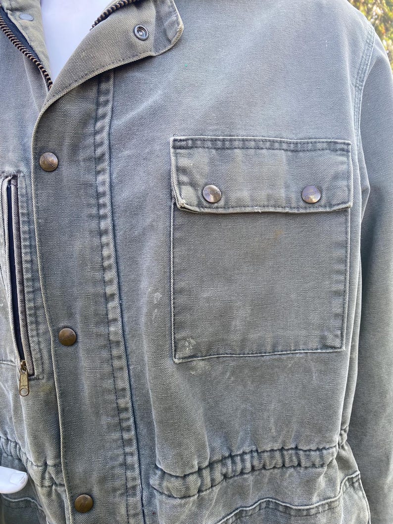 K&ouml;nnte beinhalten: Nahaufnahme einer gr&uuml;nen Canvas-Jacke mit Rei&szlig;verschluss und Druckkn&ouml;pfen. Die Jacke hat eine Brusttasche mit Druckkn&ouml;pfen und eine Taille mit Kordelzug. Der Stoff hat ein leicht abgenutztes Aussehen.