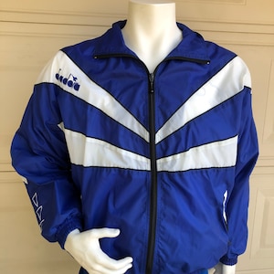 May include: A blue and white windbreaker jacket with a black zipper and the word "Dada" on the left chest. The jacket has a white and black chevron pattern.