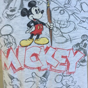 May include: A gray t-shirt with a cartoon drawing of Mickey Mouse holding a paintbrush. The shirt has the word "MICKEY" written in red across the front. The background is filled with various sketches of Mickey Mouse.