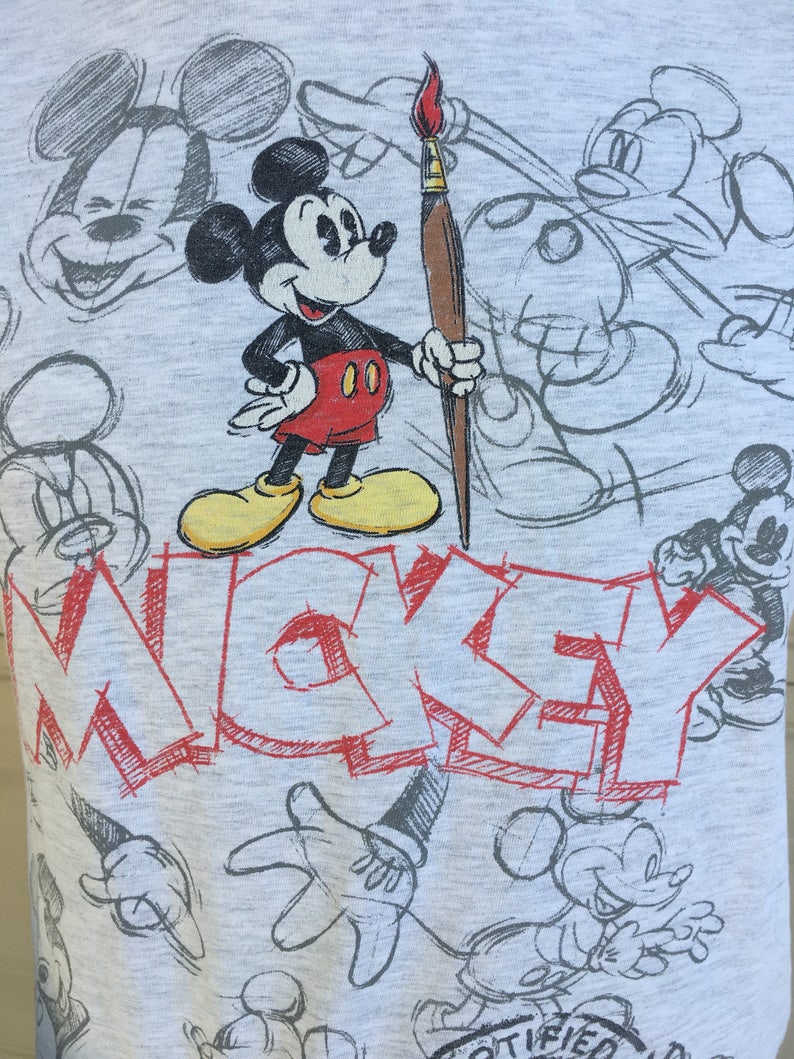 May include: A gray t-shirt with a cartoon drawing of Mickey Mouse holding a paintbrush. The shirt has the word "MICKEY" written in red across the front. The background is filled with various sketches of Mickey Mouse.