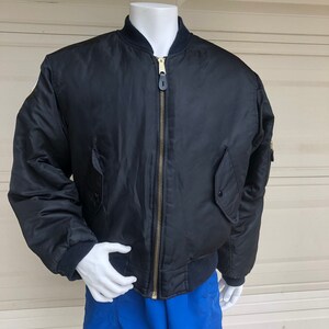 May include: A black bomber jacket with a gold zipper and a patch on the left sleeve. The jacket is on a mannequin wearing blue shorts.