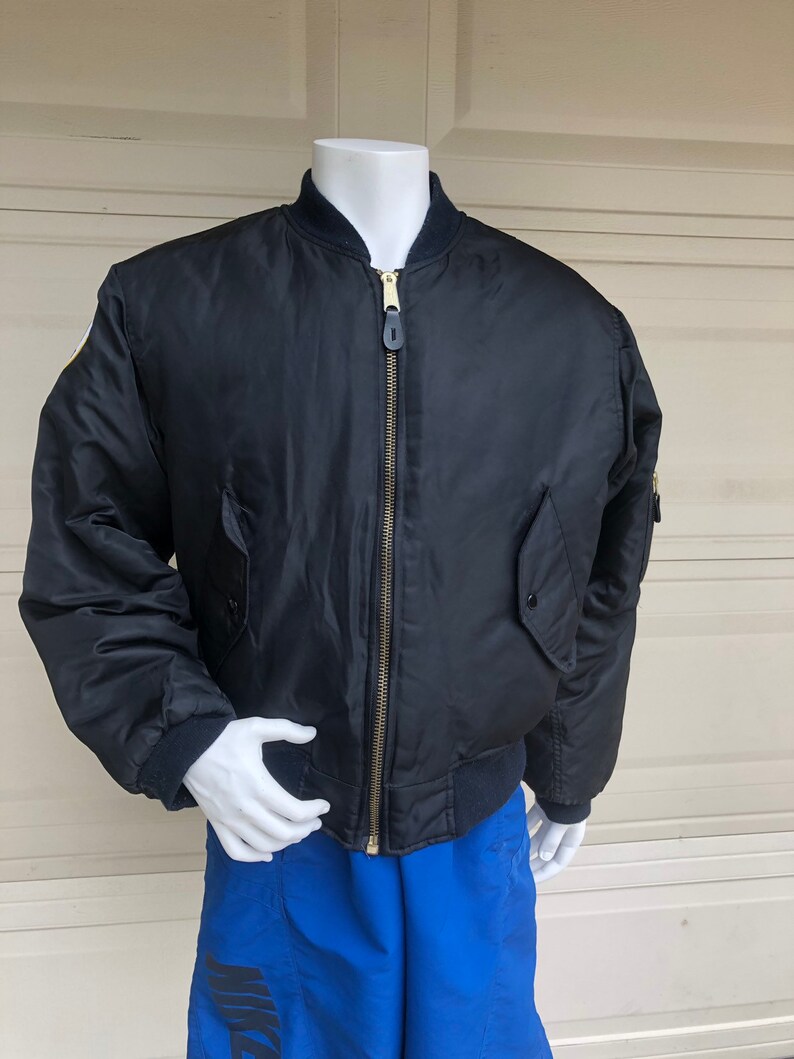 May include: A black bomber jacket with a gold zipper and a patch on the left sleeve. The jacket is on a mannequin wearing blue shorts.