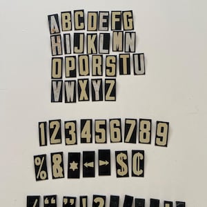 May include: A collection of vintage letter and number tiles. The tiles are black with off-white lettering and symbols. The set includes the alphabet, numbers 1-9, and various punctuation marks and symbols.