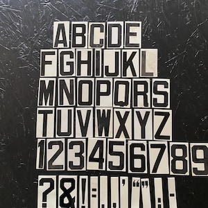 May include: A collection of black and white letter and number tiles, including the alphabet, numbers 1-9, and punctuation marks. The tiles are arranged on a dark, textured surface, ideal for crafting or educational projects.