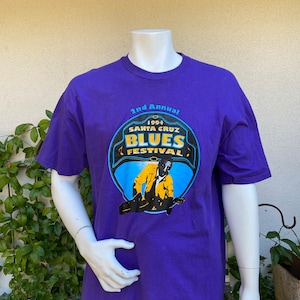 May include: Purple t-shirt with a blue and yellow graphic of a man playing a guitar. The graphic says "1994 Santa Cruz Blues Festival 2nd Annual".