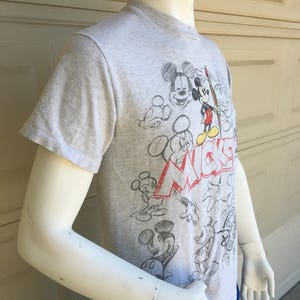 May include: A gray t-shirt with a Mickey Mouse design. The shirt features a variety of sketches of Mickey Mouse in different poses, including him holding a paintbrush. The text "Mickey" is written in red and white letters on the shirt.
