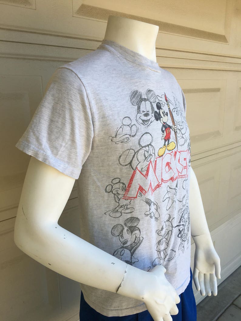 May include: A gray t-shirt with a Mickey Mouse design. The shirt features a variety of sketches of Mickey Mouse in different poses, including him holding a paintbrush. The text "Mickey" is written in red and white letters on the shirt.