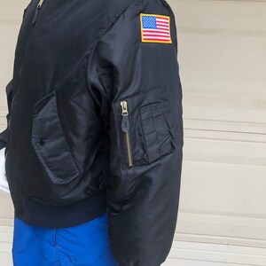 May include: A black bomber jacket with a zipper closure and a US flag patch on the sleeve. The jacket has a ribbed collar and cuffs.