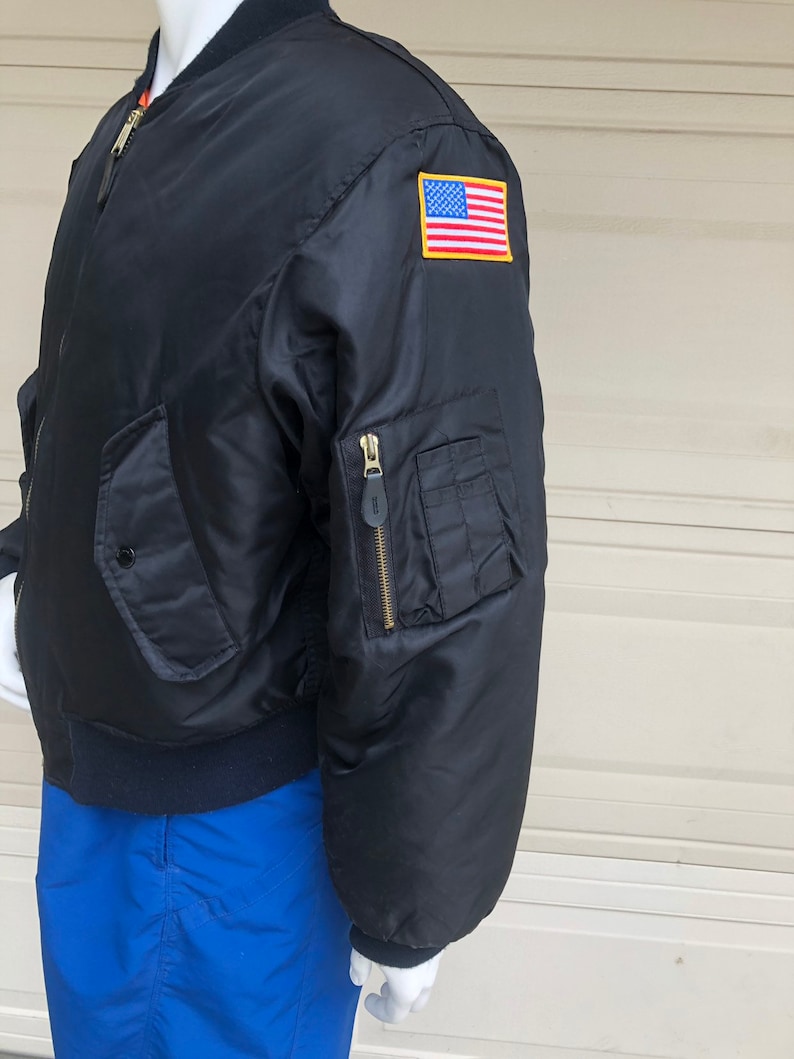 May include: A black bomber jacket with a zipper closure and a US flag patch on the sleeve. The jacket has a ribbed collar and cuffs.