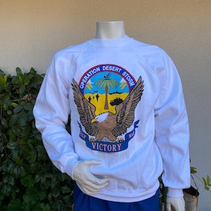 May include: White sweatshirt with a graphic print of an eagle with the text "Operation Desert Storm" and "Victory" on a blue ribbon. The eagle is holding a yellow banner with the text "Gulf War".