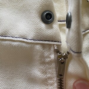May include: Close-up of a white zip with a metal zip pull that says "SCOVILL" on it. The zip is sewn into a white fabric with a blue stitching detail.