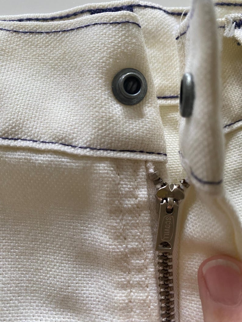 May include: Close-up of a white zip with a metal zip pull that says "SCOVILL" on it. The zip is sewn into a white fabric with a blue stitching detail.