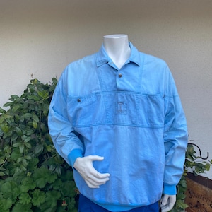 May include: A light blue long-sleeved shirt with a button-up collar and a single button closure. The shirt has a patch pocket on the chest with a small logo.