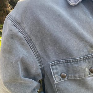 K&ouml;nnte beinhalten: Nahaufnahme einer grauen Arbeitsjacke mit einer gekn&ouml;pften Brusttasche. Die Jacke hat ein abgenutztes Aussehen mit sichtbaren N&auml;hten und zwei braunen Kn&ouml;pfen. Der Kragen ist sichtbar, und der Stoff scheint eine strapazierf&auml;hige Baumwollmischung zu sein.