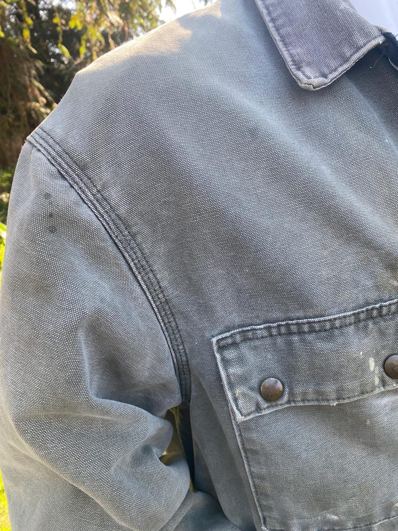 K&ouml;nnte beinhalten: Nahaufnahme einer grauen Arbeitsjacke mit einer gekn&ouml;pften Brusttasche. Die Jacke hat ein abgenutztes Aussehen mit sichtbaren N&auml;hten und zwei braunen Kn&ouml;pfen. Der Kragen ist sichtbar, und der Stoff scheint eine strapazierf&auml;hige Baumwollmischung zu sein.