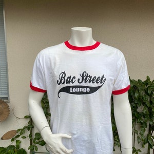 May include: White t-shirt with red trim around the collar and sleeves. The shirt features the text "Bac Street Lounge" in a black script font. The shirt is displayed on a mannequin.