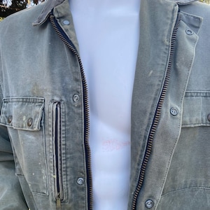 K&ouml;nnte beinhalten: Eine verblasste olivgr&uuml;ne Jacke mit Rei&szlig;verschluss und mehreren Taschen. Die Jacke hat einen Cordkragen und Knopfdetails. Das Kleidungsst&uuml;ck wird auf einer Schaufensterpuppe pr&auml;sentiert, die ihr Design und ihren Stil zeigt. Die Jacke scheint aus einem strapazierf&auml;higen, m&ouml;glicherweise leinwandartigen Material zu bestehen.