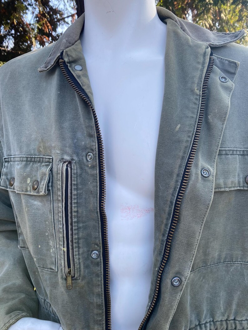 K&ouml;nnte beinhalten: Eine verblasste olivgr&uuml;ne Jacke mit Rei&szlig;verschluss und mehreren Taschen. Die Jacke hat einen Cordkragen und Knopfdetails. Das Kleidungsst&uuml;ck wird auf einer Schaufensterpuppe pr&auml;sentiert, die ihr Design und ihren Stil zeigt. Die Jacke scheint aus einem strapazierf&auml;higen, m&ouml;glicherweise leinwandartigen Material zu bestehen.