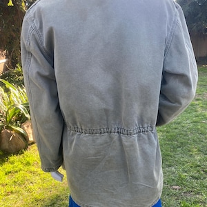 K&ouml;nnte beinhalten: Olivgr&uuml;ne Jacke mit Kordelzug in der Taille und dunkelbraunem Kragen. Die Jacke hat eine Knopfleiste und zwei Vordertaschen. Hergestellt aus strapazierf&auml;higem Material, geeignet f&uuml;r den Au&szlig;enbereich.