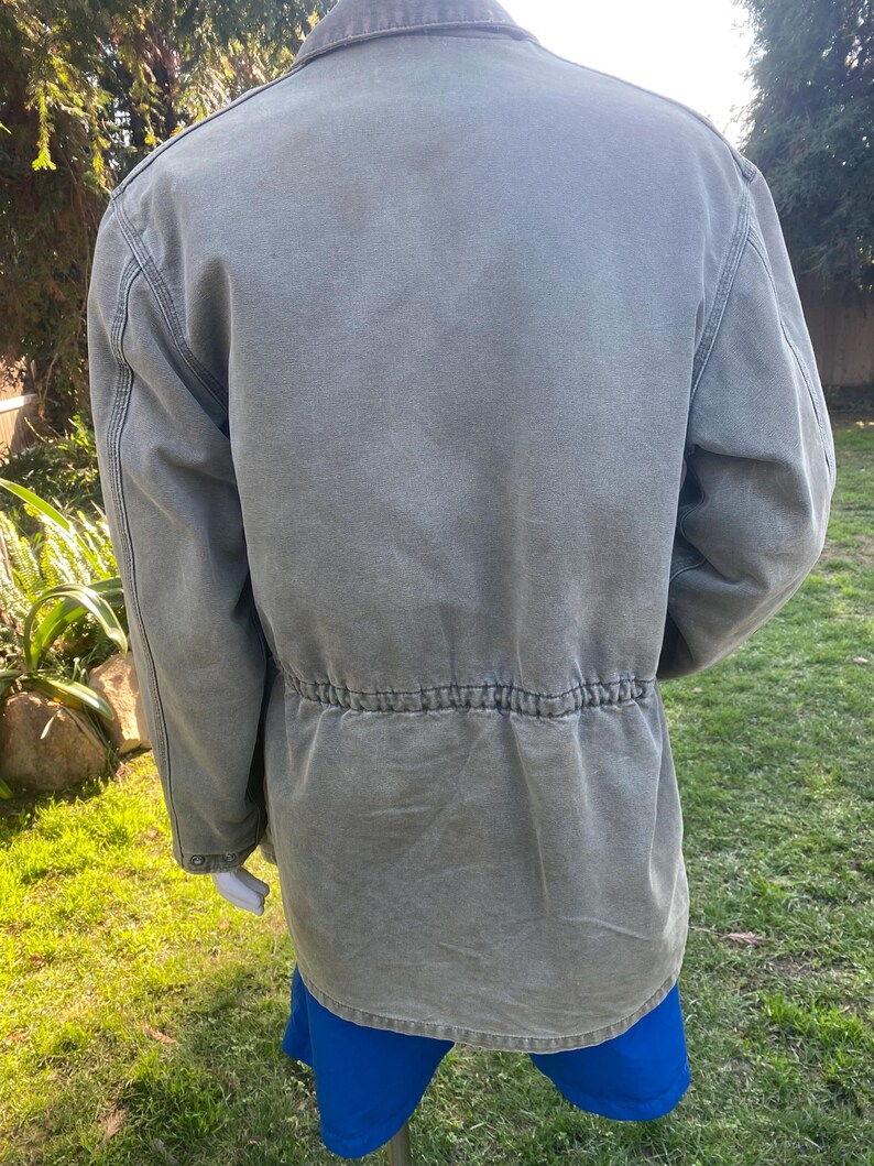 K&ouml;nnte beinhalten: Olivgr&uuml;ne Jacke mit Kordelzug in der Taille und dunkelbraunem Kragen. Die Jacke hat eine Knopfleiste und zwei Vordertaschen. Hergestellt aus strapazierf&auml;higem Material, geeignet f&uuml;r den Au&szlig;enbereich.