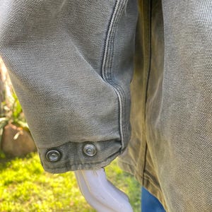 K&ouml;nnte beinhalten: Nahaufnahme eines olivgr&uuml;nen Jacken&auml;rmels. Der &Auml;rmel hat zwei silberne Druckkn&ouml;pfe am B&uuml;ndchen. Die Jacke hat sichtbare Nahtdetails und einen strukturierten Stoff. Der Hintergrund ist verschwommen, mit gr&uuml;nem Gras und einer blauen Hose.