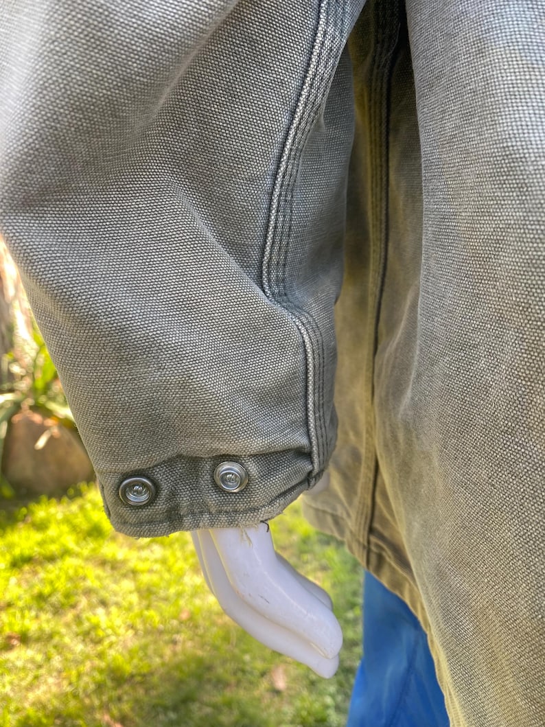 K&ouml;nnte beinhalten: Nahaufnahme eines olivgr&uuml;nen Jacken&auml;rmels. Der &Auml;rmel hat zwei silberne Druckkn&ouml;pfe am B&uuml;ndchen. Die Jacke hat sichtbare Nahtdetails und einen strukturierten Stoff. Der Hintergrund ist verschwommen, mit gr&uuml;nem Gras und einer blauen Hose.