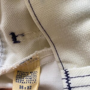May include: A white Wrangler jeans care label with blue stitching. The label has instructions for washing and drying the jeans. It also states "A product of Wrangler" and the size "36 x 32".