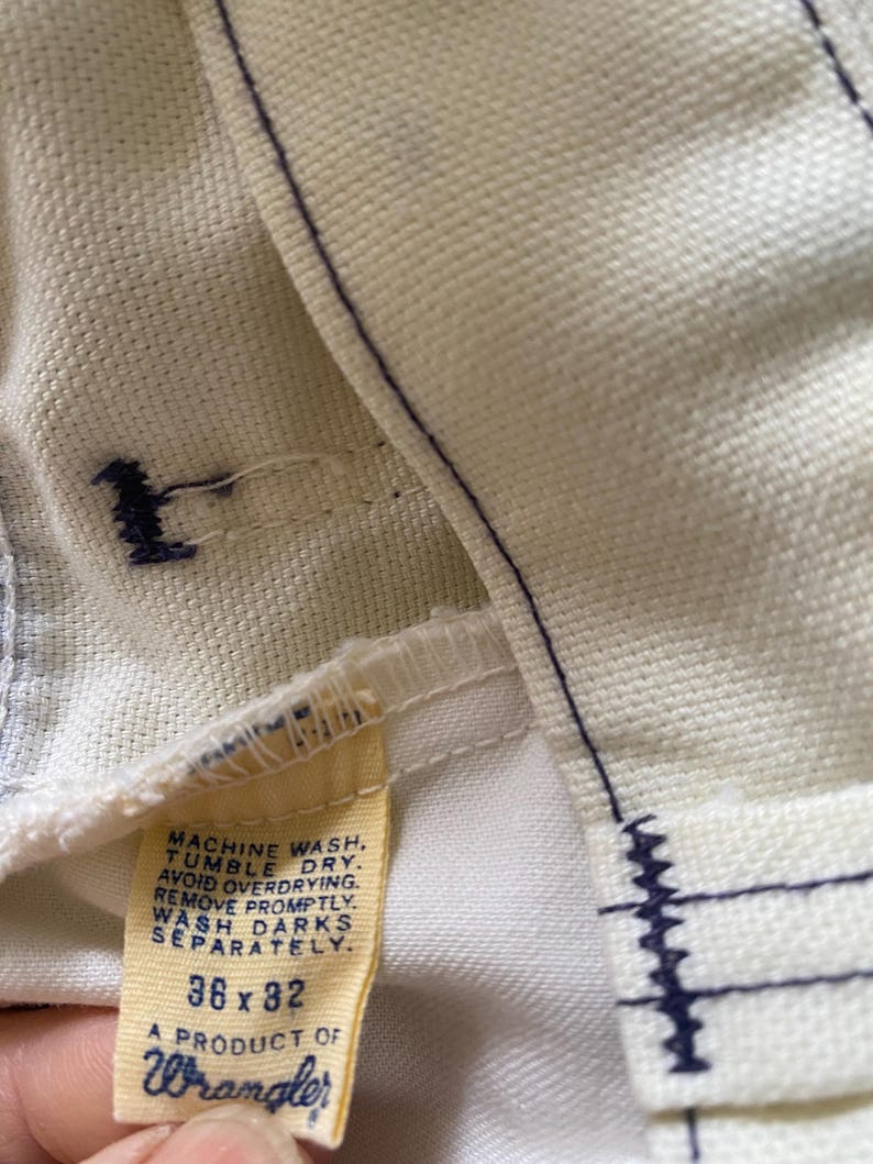 May include: A white Wrangler jeans care label with blue stitching. The label has instructions for washing and drying the jeans. It also states "A product of Wrangler" and the size "36 x 32".