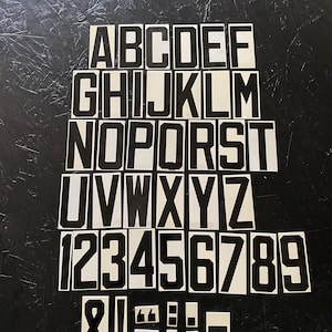 May include: Black and white alphabet and number stickers arranged on a dark surface. The stickers include the letters A through Z, numbers 1 through 9, and various punctuation marks. The font is bold and sans-serif.