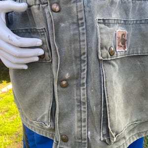 K&ouml;nnte beinhalten: Nahaufnahme einer verblichenen olivgr&uuml;nen Arbeitsjacke mit mehreren Taschen und messingfarbenen Kn&ouml;pfen. Die Jacke hat eine Taille mit Kordelzug und einen kleinen Aufn&auml;her auf einer Tasche. Die Jacke wird &uuml;ber blauen Shorts getragen.