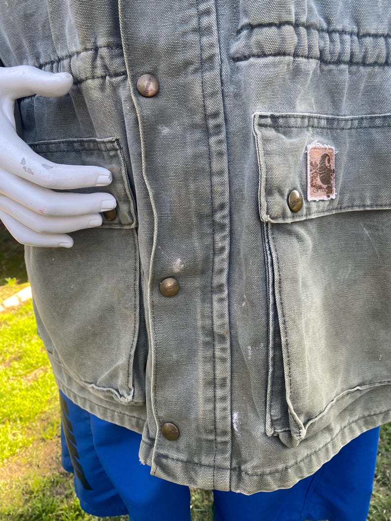 K&ouml;nnte beinhalten: Nahaufnahme einer verblichenen olivgr&uuml;nen Arbeitsjacke mit mehreren Taschen und messingfarbenen Kn&ouml;pfen. Die Jacke hat eine Taille mit Kordelzug und einen kleinen Aufn&auml;her auf einer Tasche. Die Jacke wird &uuml;ber blauen Shorts getragen.