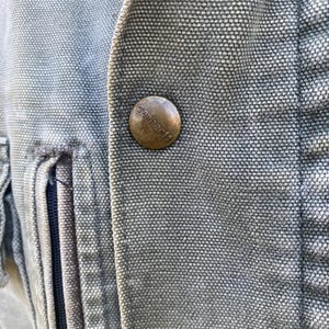 K&ouml;nnte beinhalten: Nahaufnahme einer Carhartt-Jacke. Die Jacke hat eine verwaschene blau-graue Farbe mit einem strukturierten Stoff. Ein runder, bronzefarbener Knopf mit dem Carhartt-Logo ist sichtbar. Die N&auml;hte und der Rei&szlig;verschluss der Jacke sind ebenfalls zu sehen.