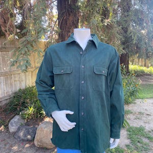 May include: A dark green long-sleeved shirt with two chest pockets and a button-down front. The shirt is displayed on a mannequin with blue jeans. The background features trees and a grassy area.