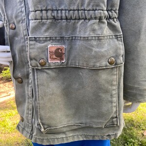 K&ouml;nnte beinhalten: Nahaufnahme einer gr&uuml;nen Carhartt-Jacke mit einer gro&szlig;en Tasche und einem kleinen Logo-Patch. Die Jacke hat einen Kordelzug in der Taille und Metallkn&ouml;pfe. Der Stoff wirkt abgenutzt, mit einer Vintage-&Auml;sthetik.