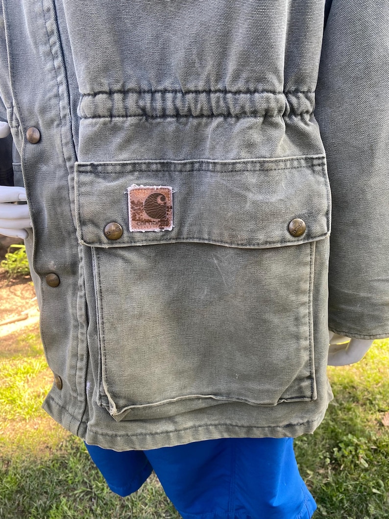 K&ouml;nnte beinhalten: Nahaufnahme einer gr&uuml;nen Carhartt-Jacke mit einer gro&szlig;en Tasche und einem kleinen Logo-Patch. Die Jacke hat einen Kordelzug in der Taille und Metallkn&ouml;pfe. Der Stoff wirkt abgenutzt, mit einer Vintage-&Auml;sthetik.
