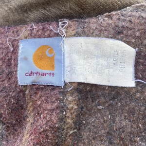 K&ouml;nnte beinhalten: Nahaufnahme eines Carhartt-Etiketts auf einem braunen und weinroten Stoff. Das Etikett ist hellblau mit dem Carhartt-Logo in Orange und dem Markennamen in Rot. Ein wei&szlig;es Etikett mit Text ist angebracht.