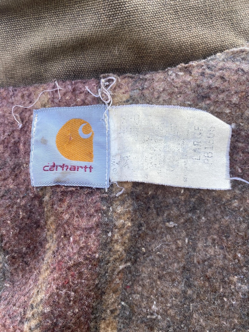 K&ouml;nnte beinhalten: Nahaufnahme eines Carhartt-Etiketts auf einem braunen und weinroten Stoff. Das Etikett ist hellblau mit dem Carhartt-Logo in Orange und dem Markennamen in Rot. Ein wei&szlig;es Etikett mit Text ist angebracht.