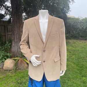 May include: A tan corduroy blazer with two buttons and a notched lapel is displayed on a mannequin. The mannequin is also wearing blue swim trunks. The blazer has a chest pocket and two lower pockets. The background is a grassy yard with trees.