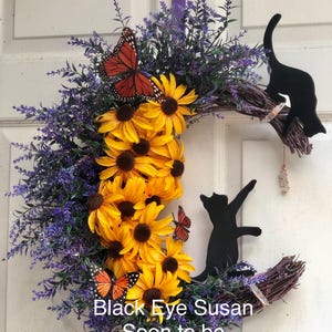 May include: A crescent moon-shaped wreath with a black cat silhouette, purple lavender, and yellow sunflowers. There are also orange and black butterfly accents. The wreath has the text "Black Eye Susan Soon to be Discontinued".